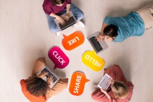 Vier Personen, die mit Laptops und Tablets im Kreis auf dem Boden sitzen, umgeben von bunten Sprechblasen mit den Aufschriften KLICK, LIKE, HELLO und SHARE.
